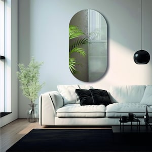 May include: A white sofa with black cushions in front of a wall with a large oval mirror. The mirror is reflecting a plant with green leaves.