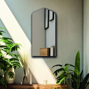May include: A wall-mounted mirror with a pointed arch top and a flat bottom edge. The reflective surface shows a partial view of a room interior, including a wooden block and framed wall art, creating a clear reflection of the surrounding space.