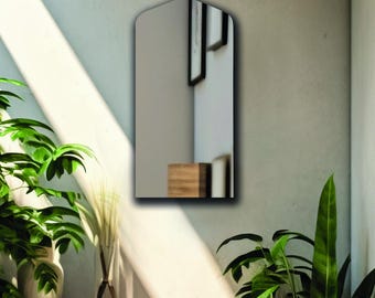 May include: A wall-mounted mirror with a pointed arch top and a flat bottom edge. The reflective surface shows a partial view of a room interior, including a wooden block and framed wall art, creating a clear reflection of the surrounding space.