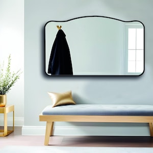 Peut inclure: Un miroir arqué encadré de noir avec un manteau suspendu à un crochet. Le miroir est suspendu à un mur bleu clair au-dessus d'un banc rembourré gris avec un coussin doré. Le banc a une structure en bois clair.