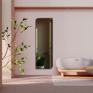 May include: A tall, rectangular wall mirror with a rounded top corner on the left side. The mirror features a reflective surface that shows a partial view of an indoor space, including a potted plant and light streaming through a window.
