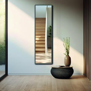 Puede incluir: Un espejo de cuerpo entero con marco negro, que refleja una escalera de madera y una planta en maceta. El espejo está situado en una habitación con suelo de madera y un puf de tejido negro.