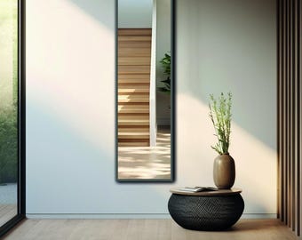 May include: A tall, rectangular wall mirror with a dark frame. The mirror reflects a wooden staircase and a green plant. Beside the mirror sits a round, black textured side table holding a ceramic vase with tall green stems and leaves.