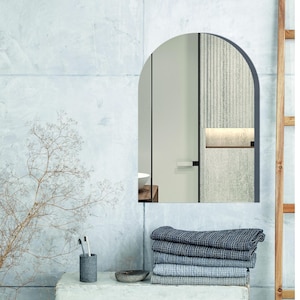 May include: An arched wall mirror with a thin dark frame. The mirror reflects a room with a door, a light-colored wall, and a shelf. Below the mirror sits a stack of five folded blue and gray textured towels, a gray cup with toothbrushes, and a small bowl.