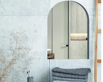 May include: An arched wall mirror with a thin dark frame. The mirror reflects a room with a door, a light-colored wall, and a shelf. Below the mirror sits a stack of five folded blue and gray textured towels, a gray cup with toothbrushes, and a small bowl.