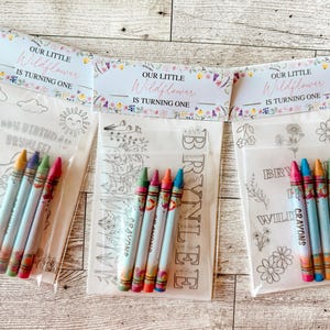 May include: Three clear plastic bags with crayons and coloring pages. Each bag has a header that reads "OUR LITTLE Wildflower IS TURNING ONE." The coloring pages have various designs and the crayons are in multiple colors.