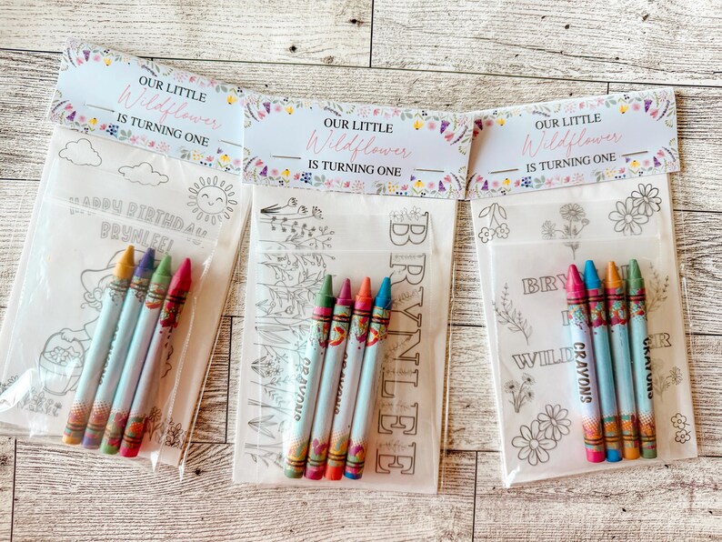 May include: Three clear plastic bags with crayons and coloring pages. Each bag has a header that reads "OUR LITTLE Wildflower IS TURNING ONE." The coloring pages have various designs and the crayons are in multiple colors.