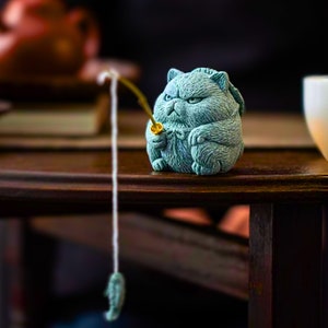 Può includere: Una statuetta di gatto in ceramica blu turchese che tiene una canna da pesca con un pesce dorato all'estremità della lenza. Il gatto è seduto su un tavolo di legno marrone scuro.
