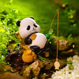 May include: A small, white and black panda figurine sitting on a miniature bamboo chair, holding a fishing rod with a green lure. The panda is wearing a black ribbon around its neck. The figurine is sitting on a bed of green moss with white pebbles in the foreground.