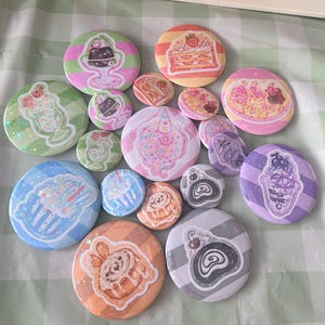 Puede incluir: Una colección de chapas redondas con diversas ilustraciones de postres, incluyendo pasteles, parfaits y repostería. Las chapas muestran fondos coloridos con rayas y patrones, exhibiendo una variedad de dulces en diferentes tamaños.