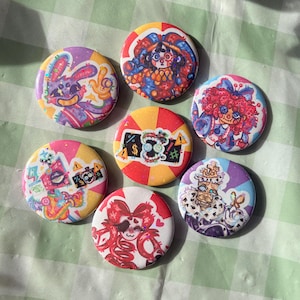 Può includere: Una collezione di sette bottoni rotondi e colorati con illustrazioni uniche. I disegni includono un coniglio, un clown e altri personaggi stravaganti. I bottoni sono disposti su una superficie a scacchi verdi e bianchi.