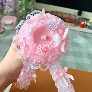 Op de afbeelding: Een ronde roze rozet met lagen van geplooide stof, witte kanten randen en satijnen linten. Het midden is versierd met kleine stoffen roosjes, een witte bedel in de vorm van een botje en een miniatuur blauw-witte blisterverpakking.