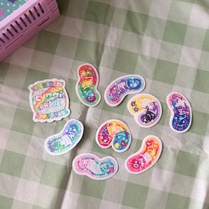 May include: A selection of colourful stickers in various shapes, including jelly beans. The stickers feature vibrant designs with rainbows, stars, and the text "Gummy Pride". They are arranged on a green and white checkered surface.
