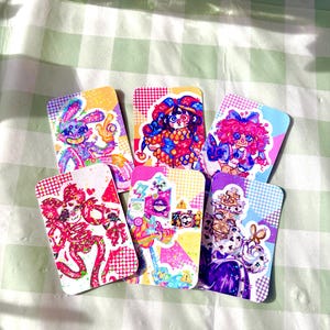 Puede incluir: Seis tarjetas artísticas rectangulares y coloridas con ilustraciones caprichosas. Las tarjetas presentan varios personajes, incluyendo un conejo y payasos, con colores vibrantes y detalles brillantes. Están dispuestas sobre una superficie a cuadros verdes y blancos.
