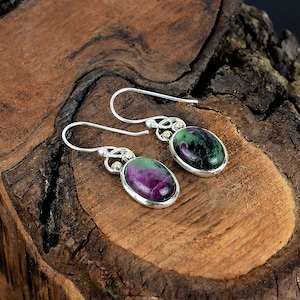 Op de afbeelding: Een paar zilveren oorbellen met ovale, groen-paarse edelstenen. De oorbellen hebben een Keltisch knoopontwerp.