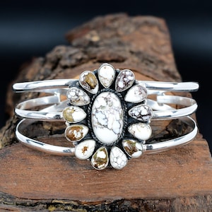 May include: A silver cuff bracelet featuring a central oval white stone with brown veining, surrounded by teardrop-shaped stones in similar colors. The bracelet has three parallel silver bands.
