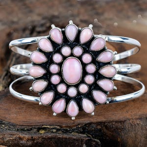 May include: A silver cuff bracelet with a central floral design. It features pink, teardrop-shaped stones and three parallel bands. The floral centerpiece has a dark, textured background.