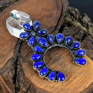 Puede incluir: Un colgante plateado con forma de media luna, adornado con numerosas piedras preciosas de color azul oscuro en forma de lágrima. El colgante se encuentra sobre un fondo de madera natural, resaltando el contraste entre el metal y las vibrantes piedras azules.