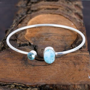 Puede incluir: Un brazalete de plata con una piedra ovalada de color azul claro y una piedra redonda más pequeña de color azul claro.