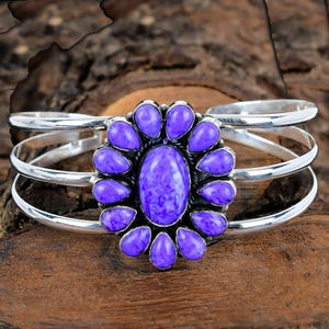 Pode incluir: Uma pulseira de punho prateada com uma pedra central oval roxa rodeada por pedras roxas em forma de gota. A pulseira tem três faixas prateadas paralelas. A joia está sobre um fundo de madeira.