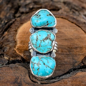 May include: A silver ring featuring three large, irregular turquoise stones. The stones are a vibrant blue-green color with dark veining. The ring has a wide band and decorative silver accents. The ring is displayed on a wooden surface.