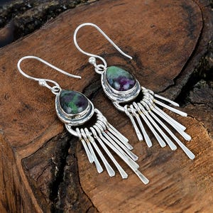 Op de afbeelding: Zilveren oorhangers met druppelvormige edelstenen in groen en paars. De oorbellen hebben een franje van zilveren staven die onder de edelsteen setting hangen. De oorbellen worden gepresenteerd op een houten oppervlak.