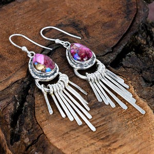 May include: A pair of silver dangle earrings with teardrop-shaped, multi-colored stones. The stones are shades of pink, purple, and gold. Below the stones are multiple silver bars, creating a fringe effect. The earrings are displayed on a wooden surface.