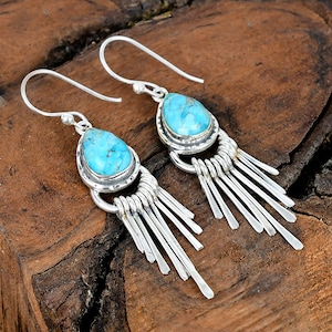 May include: A pair of silver earrings with turquoise teardrop-shaped stones. The earrings have a fringe of silver bars hanging below the stone. The earrings are displayed on a wooden surface.