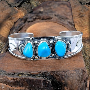 Könnte beinhalten: Silbernes Armreif-Armband mit drei leuchtenden Türkissteinen. Das Armband hat ein detailliertes Design mit einem seilartigen Rand um jeden Stein und Blattakzenten an den Seiten. Die Türkissteine sind ein leuchtendes Blau.