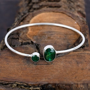 May include: A silver bangle bracelet with two green oval-shaped gemstones. The bracelet is open and has a simple design.