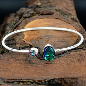 Può includere: Un bracciale a polsino argentato con due pietre preziose iridescenti. Una pietra presenta tonalità viola, verdi e blu, l'altra un verde e blu vibranti. Il bracciale è posto su uno sfondo di legno rustico. Circonferenza di circa 17 cm.