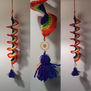 Op de afbeelding: Een handgemaakte, regenboogkleurige gehaakte spiraalvormige hangdecoratie. De spiraal is gemaakt van rood, oranje, geel, groen, blauw en paars garen. Een houten label met tekst en een kwastje zijn bevestigd. Opgehangen aan een rood touwtje.
