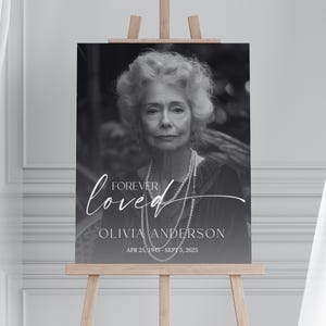 May include: A black and white memorial sign on a wooden easel. The sign features a portrait of an older woman, with the words "Forever Loved" and the name "Olivia Anderson" in white script. Dates of birth and death are also included.