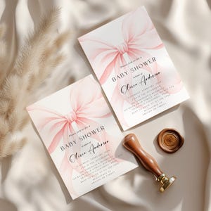 May include: Two baby shower invitations with a watercolor pink bow design. The invitations include the text "Baby Shower" and the name "Olivia Anderson". A wooden stamp and wax seal are also present.
