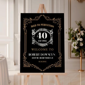 May include: A black and gold birthday welcome sign on a wooden easel. The sign reads "Aged to Perfection" above a large "40" in a decorative frame. Below, it says "Welcome to Robert Downey's 40th Birthday".