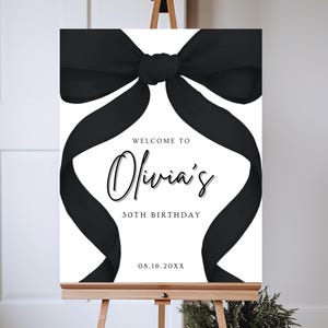 May include: A white welcome sign with a black bow on top. The sign reads "Welcome to Olivia's 30th Birthday 08.16.20XX".