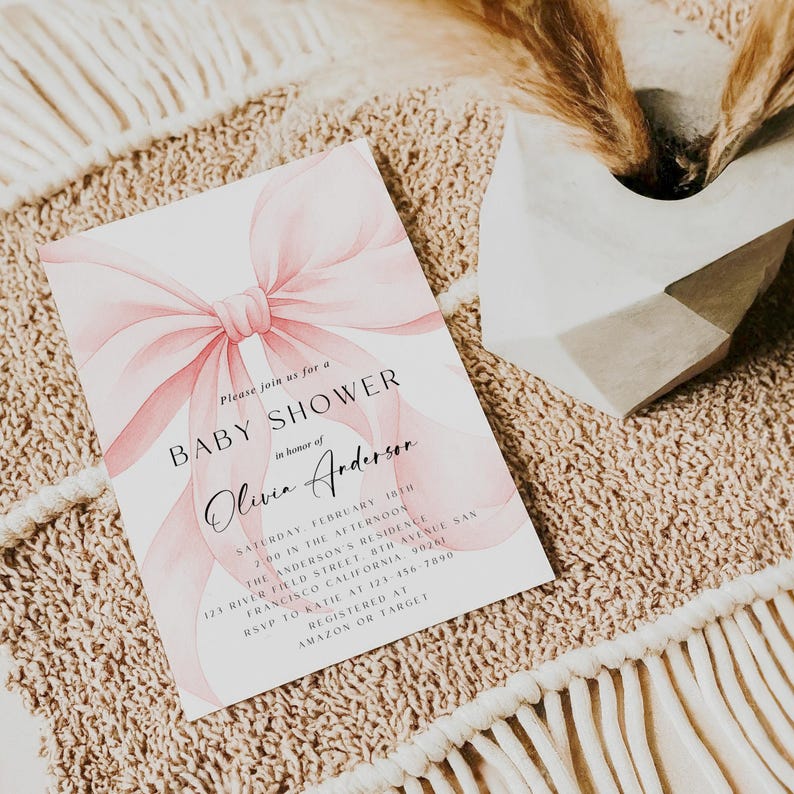 May include: A baby shower invitation with a watercolor pink bow design. The invitation includes the text "BABY SHOWER" and details for the event, including the honoree's name, date, time, and location. RSVP information and registry details are also included.