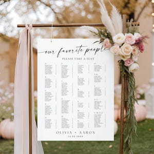 May include: A white seating chart with the text "our favorite people" and "please take a seat". The chart is decorated with flowers and ribbons, and includes names and table assignments for a wedding. The names Olivia and Aaron are listed.
