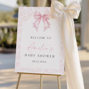 May include: A baby shower welcome sign with a pink bow illustration and the text "Welcome to Amelia's Baby Shower 08.12.20XX". The sign is on a gold easel and has a large cream-colored bow attached.