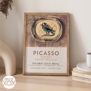 May include: Framed art print featuring a Picasso ceramic design. The artwork showcases a bird on a plate with blue, green, and yellow hues. The print includes text: "PICASSO" and "GALERIE LUCIE WEILL". The frame is made of light brown wood.