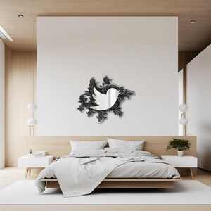 Op de afbeelding: Een zwarte, sierlijke spiegel in de vorm van een vogel met een witte achtergrond. De spiegel hangt aan een witte muur boven een bed met witte beddengoed. De kamer is minimalistisch ingericht met een lichte houten vloer en witte muren.