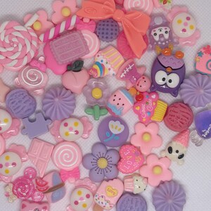 May include: A collection of colorful resin flatback cabochons featuring various cute designs, including flowers, hearts, animals, and food. The cabochons are in shades of pink, purple, and white.