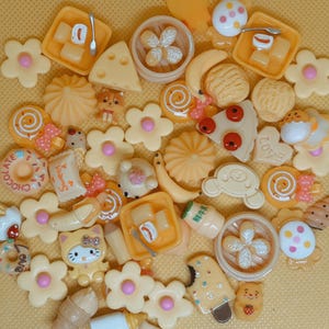 May include: A collection of miniature food-themed charms in shades of yellow, cream, and pink. The charms include a slice of cheese, a dim sum, a slice of pizza, a donut, a Hello Kitty figure, and various flowers.