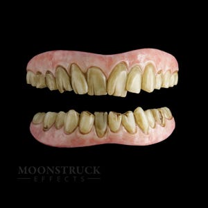 Peut inclure: Prothèse dentaire supérieure et inférieure avec des dents jaunies et tachées. Les gencives sont roses pâles. Les dents ont une apparence usée. Le fond est noir. Le texte "MOONSTRUCK EFFECTS" est en bas.