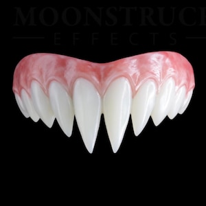 May include: A set of white vampire fangs with a pink gum line, featuring pointed teeth and a curved shape. The text MOONSTRUCK EFFECTS is visible in the background. The fangs are designed to fit over teeth and have a glossy, realistic finish.