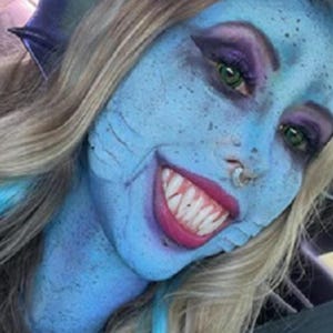 May include: A woman with blue face paint and a nose ring is smiling. Her eyes are green and she has long blonde hair.