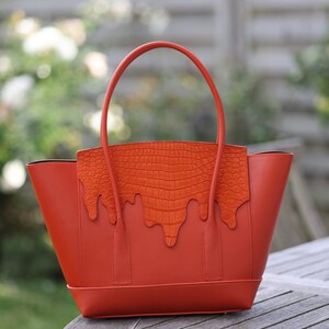 May include: A burnt orange tote bag with a crocodile-textured accent. The bag has a structured shape with a rounded top handle. The crocodile texture is a lighter shade of orange, creating a contrast. The bag is sitting on a wooden table.