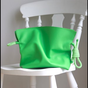 May include: A bright green leather pouch bag with rope handles, resting on a white wooden chair. The bag has a soft, folded appearance and a simple design. The rope handles are a lighter shade of green.