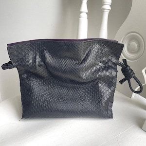 Peut inclure: Un sac pochette gris foncé avec un motif peau de serpent. Le sac a une fermeture à cordon avec un détail noué et un intérieur violet. Le sac est posé sur une surface blanche.