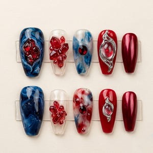 May include: A collection of ten artificial nails with various designs. The nails feature a range of colors, including deep blue, clear, red, and metallic burgundy. Some nails have embedded red crystal-like embellishments, while others showcase intricate patterns and silver accents.